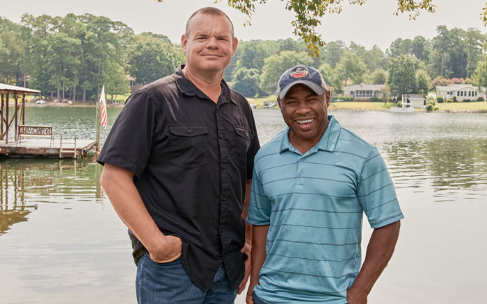 John and Darrell (wearing baseball hat) smiling. Water and dock in the background